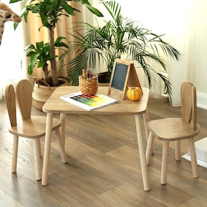 May include: A light wood children's table and two chairs. The chairs have bunny ear-shaped backs. A small chalkboard, a pumpkin, and a basket of pencils sit on the table. A color palette and a variety of plants are in the background.