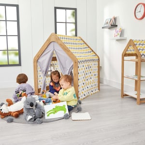 Op de afbeelding: Een houten speelhuisje met een dak en muren van gele en grijze stof met sterrenpatroon. Kinderen spelen in en rond het speelhuisje. Een grijs tapijt met pompons en knuffels staat op de voorgrond. Een houten boekenrek staat rechts.