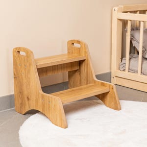 Puede incluir: Un taburete de madera de dos escalones con acabado natural. El taburete tiene forma rectangular con bordes redondeados y asas recortadas en los laterales. Los escalones son anchos y resistentes, diseñados para uso infantil. Una pequeña cuna de madera en el fondo.