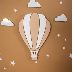 May include: A decorative wall art piece featuring a hot air balloon in beige and white, surrounded by white clouds and stars. The artwork is designed to be mounted on a brown wall, creating a whimsical nursery or playroom theme.