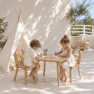 Op de afbeelding: Een kindertafel van lichtgekleurd hout met twee bijpassende stoeltjes met gebogen rugleuningen in de vorm van een gewei. Het meubilair staat op een terras naast een witte stoffen tipi, waar twee kinderen op wit papier aan het tekenen zijn.