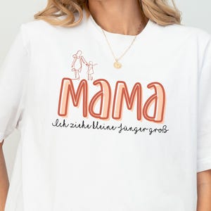 Könnte beinhalten: Weißes Oversize-T-Shirt mit einer Grafik einer Mutter und eines Kindes, die sich an den Händen halten. Der Text "Mama" ist in großen, bunten Buchstaben mit dem Satz "ich ziehe kleine finger groß" darunter.