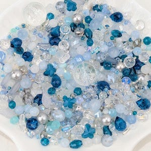 May include: A selection of blue and clear beads in a white, scalloped dish. The beads are of various shapes, including round, teardrop, and flower-shaped, and range in colour from light to dark blue, with some silver accents. The dish is white.