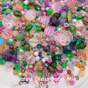 Könnte beinhalten: Eine Mischung aus Glasperlen in verschiedenen Formen und Farben, darunter Rosa, Grün, Lila und transparent. Die Perlen liegen auf einer weißen Oberfläche. Der Text lautet "Curated Glass Bead Mix" mit Größenoptionen.