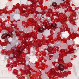 May include: A selection of red, white, and clear beads in diverse shapes and sizes. The beads include round, oval, and faceted designs, with some shaped like flowers and strawberries. The beads are scattered on a white surface.