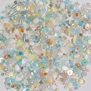 May include: A collection of assorted beads in shades of blue, white, yellow, and peach, including pearls, starfish, and heart-shaped beads. The beads are scattered on a white, scalloped-edge surface. The text "CELESTIA BEADS & CRAFTS" is visible.