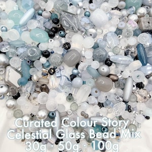 May include: A collection of glass beads in a range of shapes and sizes, in white, grey, and blue hues. The beads include round, oval, and faceted designs. The text "Curated Colour Story Celestial Glass Bead Mix" is visible.