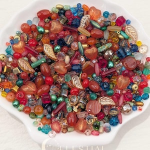 May include: A collection of assorted loose beads in various shapes, sizes, and colours, including red, orange, blue, green, and metallic gold tones. The beads feature heart, leaf, and cylindrical forms. The text CELESTIAL BEADS & CRAFTS is visible.