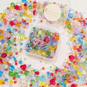 May include: A vibrant assortment of multi-coloured beads in a clear plastic container, with many more scattered around. The beads are in various shapes, including hearts and squares. The container has a sticker that reads "Celestial Beads & Crafts".