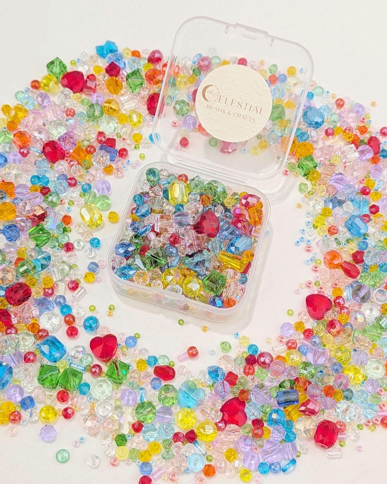 May include: A vibrant assortment of multi-coloured beads in a clear plastic container, with many more scattered around. The beads are in various shapes, including hearts and squares. The container has a sticker that reads "Celestial Beads & Crafts".