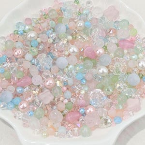 Peut inclure: Un assortiment de perles et d'ornements aux couleurs pastel, de formes variées, notamment rondes, florales et à facettes. Les perles sont dans des tons de rose, de bleu, de vert et de transparent, créant une esthétique délicate et fantaisiste.