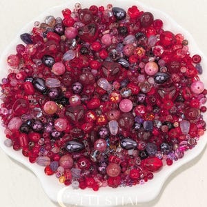 May include: A collection of assorted beads in shades of red, pink, and dark purple, featuring various shapes including spheres, teardrops, and hearts, contained within a white scalloped dish with the text CELESTIAL BEADS & CRAFTS printed at the bottom.