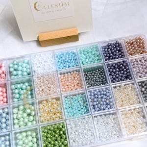 Peut inclure: Un organisateur en plastique transparent rempli de perles de diff&eacute;rentes couleurs. Les perles sont dans des tons de rose, bleu, vert, blanc et or. Le texte "Celestial Beads & Crafts" est visible sur une carte en arri&egrave;re-plan. Le texte "Beads not included, for demo only" est en bas.