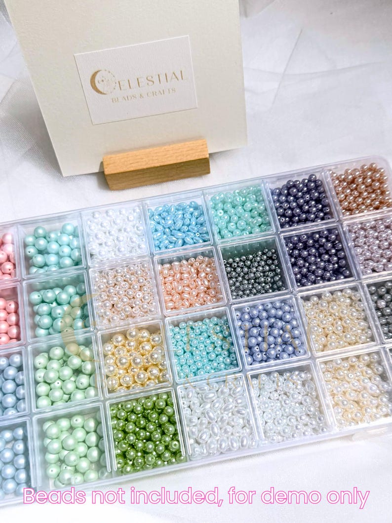 Peut inclure: Un organisateur en plastique transparent rempli de perles de diff&eacute;rentes couleurs. Les perles sont dans des tons de rose, bleu, vert, blanc et or. Le texte "Celestial Beads & Crafts" est visible sur une carte en arri&egrave;re-plan. Le texte "Beads not included, for demo only" est en bas.