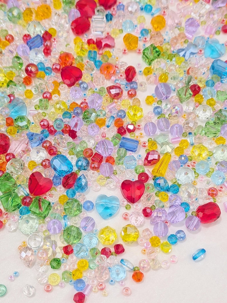 May include: A colourful collection of beads in diverse shapes and sizes. The beads include heart-shaped, round, and faceted designs in shades of red, blue, yellow, green, and purple. These beads are suitable for jewellery making and craft projects.