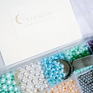 Peut inclure: Un organisateur en plastique transparent rempli de diverses perles color&eacute;es. Les perles sont dans des tons de blanc, de bleu, de vert et de p&ecirc;che. Une paire de ciseaux argent&eacute;s repose dans l'organisateur. Le texte "CELESTIAL BEADS & CRAFTS" est visible.