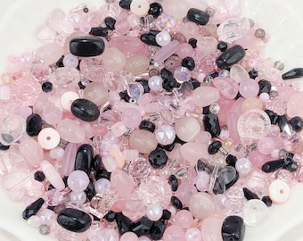 May include: A collection of assorted loose beads in various shades of pink, clear, and black. The beads feature diverse shapes, including round, oval, faceted, and butterfly-like forms, with varying textures ranging from smooth and opaque to translucent and sparkling.