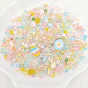 May include: A selection of pastel beads and charms in a white, shell-shaped dish. The beads are in shades of pink, yellow, blue, and iridescent colours. The assortment includes Easter-themed charms, such as an egg and a bunny.
