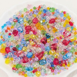 May include: A colourful collection of beads in a variety of shapes and sizes, including round, faceted, and star-shaped. The beads are a mix of red, pink, yellow, blue, green, and clear, presented in a white, shallow dish. Perfect for jewellery making.