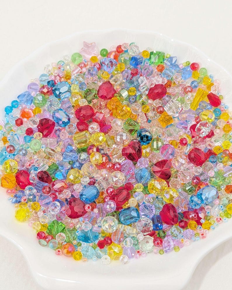 May include: A colourful collection of beads in a variety of shapes and sizes, including round, faceted, and star-shaped. The beads are a mix of red, pink, yellow, blue, green, and clear, presented in a white, shallow dish. Perfect for jewellery making.