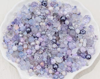 May include: A collection of assorted beads in shades of lavender, periwinkle, light blue, and white, featuring various shapes including round, faceted, floral, and teardrop styles, displayed on a white scalloped dish with the text CELESTIAL BEADS & CRAFTS.