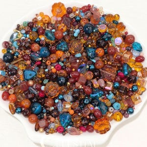 May include: A large collection of assorted loose beads in a white scalloped dish. The beads vary in size, shape, and texture, featuring a colour palette of amber, orange, deep red, turquoise, teal, and dark blue, with some translucent finishes.