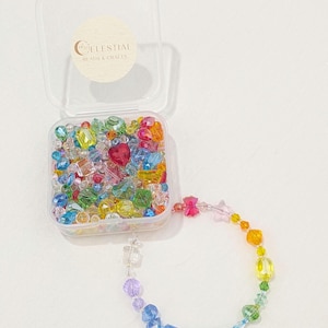 May include: A clear plastic container filled with colourful beads, including heart-shaped and star-shaped beads. A rainbow-coloured beaded bracelet is attached to the container. The container has a sticker that reads "CELESTIAL BEADS & CRAFTS."