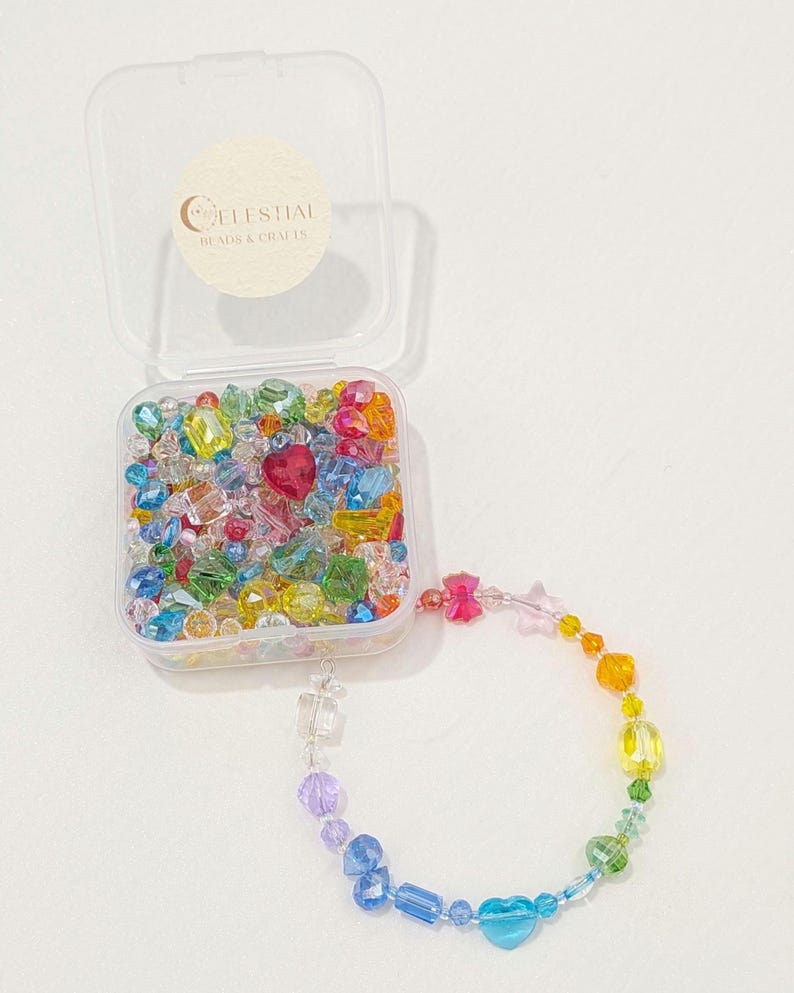 May include: A clear plastic container filled with colourful beads, including heart-shaped and star-shaped beads. A rainbow-coloured beaded bracelet is attached to the container. The container has a sticker that reads "CELESTIAL BEADS & CRAFTS."