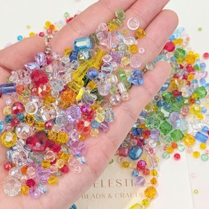 May include: A hand displaying a collection of colourful beads in various shapes and sizes. The beads are in shades of red, blue, yellow, and green, with clear and opaque finishes. The text "CELESTIA BEADS & CRAFTS" is visible.
