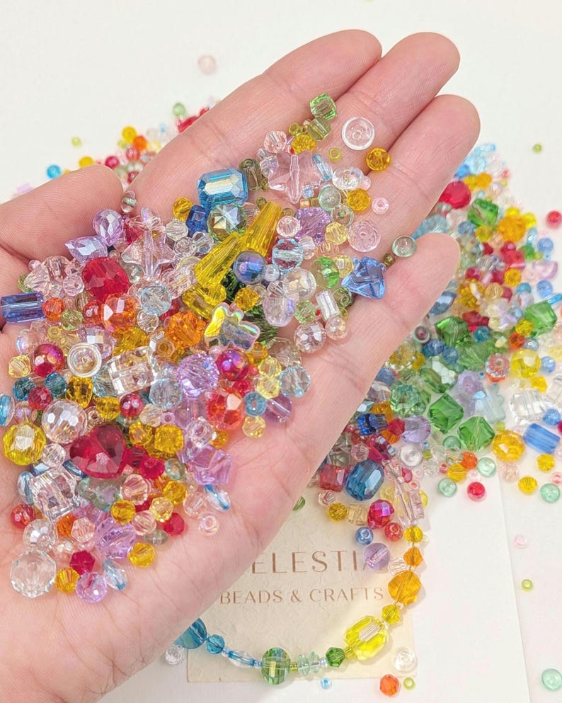 May include: A hand displaying a collection of colourful beads in various shapes and sizes. The beads are in shades of red, blue, yellow, and green, with clear and opaque finishes. The text "CELESTIA BEADS & CRAFTS" is visible.