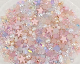 May include: A collection of pastel-colored beads in a white, scalloped dish. The beads include various shapes and sizes, such as round, star, and faceted, in shades of pink, white, and iridescent. These beads are ideal for jewelry making and crafting.