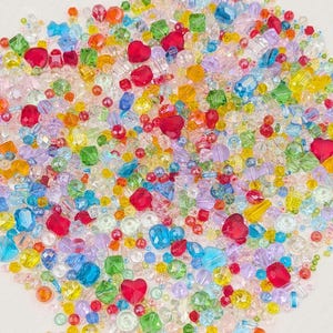 May include: A colourful assortment of beads in various shapes and sizes. The mix includes heart-shaped, square, and round beads in shades of red, blue, green, yellow, and purple. These beads are ideal for jewellery making and crafting projects.
