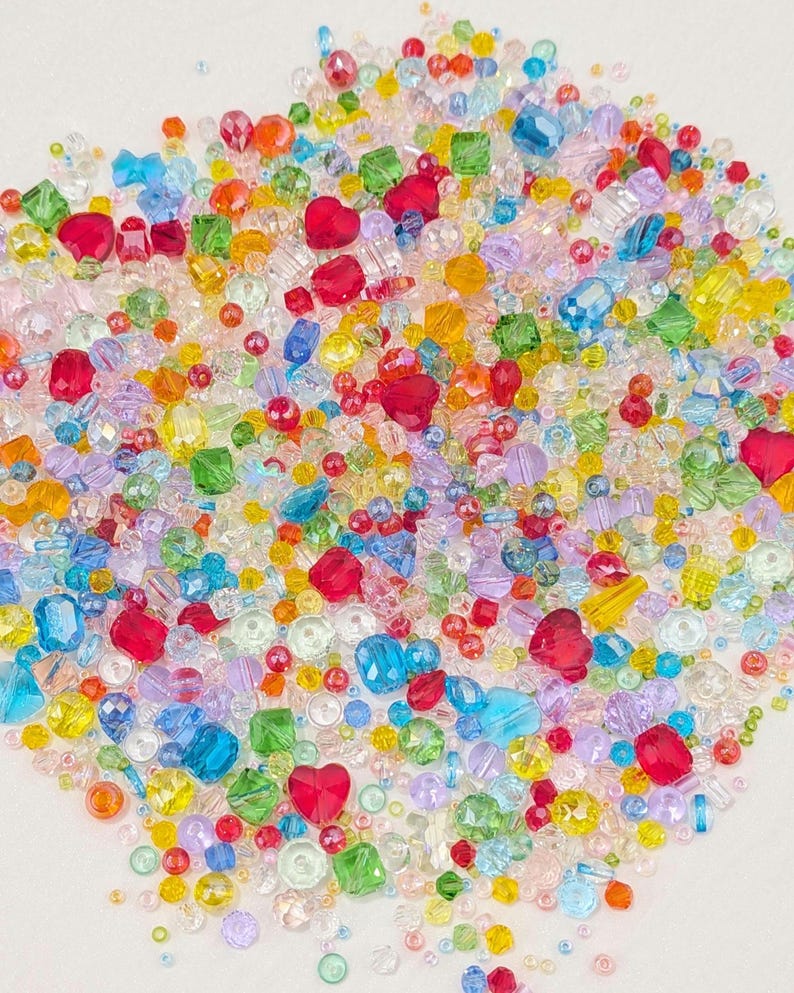May include: A colourful assortment of beads in various shapes and sizes. The mix includes heart-shaped, square, and round beads in shades of red, blue, green, yellow, and purple. These beads are ideal for jewellery making and crafting projects.