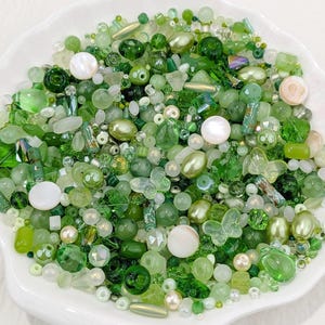 Op de afbeelding: Een verzameling groene kralen in verschillende vormen en maten, waaronder rond, ovaal en vlindervormig, gepresenteerd in een wit, geschulpte schaal. De kralen variëren in tinten van licht- tot donkergroen, met iriserende en parelachtige afwerkingen.