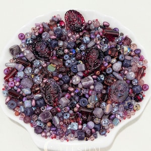 May include: A collection of assorted beads in shades of purple, violet, and plum, including faceted crystals, smooth round spheres, and several large, dark purple oval beads featuring a raised rose floral design, all contained in a white dish.