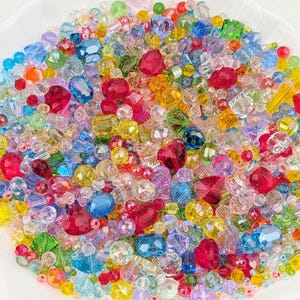 May include: A colourful assortment of beads in various shapes and sizes, including faceted crystals, round beads, and heart-shaped beads. The beads are in shades of red, yellow, blue, green, and purple, creating a rainbow effect. The beads are displayed in a white, scalloped dish.