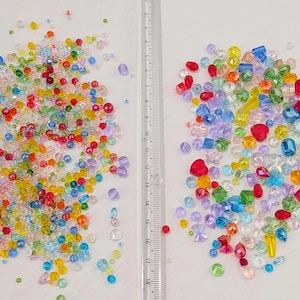 May include: A selection of colourful beads in various shapes and sizes, including round, heart, and faceted beads. The beads are in shades of red, yellow, green, blue, and purple. A ruler is placed between the two piles of beads.