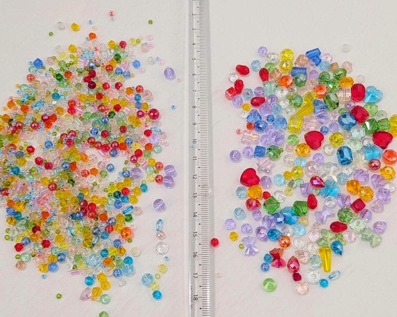 May include: A selection of colourful beads in various shapes and sizes, including round, heart, and faceted beads. The beads are in shades of red, yellow, green, blue, and purple. A ruler is placed between the two piles of beads.