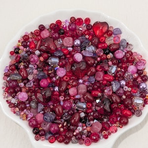 May include: A collection of assorted beads in various shades of red, pink, and purple, including round, faceted, and heart-shaped glass and plastic pieces, all contained within a white scalloped-edge dish for crafting and jewellery making.
