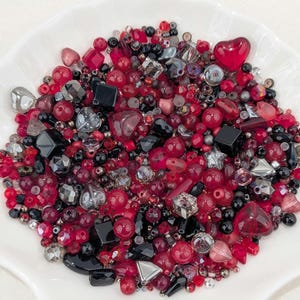 May include: A mix of red, black, and silver beads in various shapes and sizes, including hearts, cubes, and spheres, are arranged in a white dish. The beads are made of glass and other materials, creating a textured and reflective surface.
