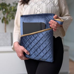 May include: A blue velvet clutch with a quilted pattern and gold trim. The clutch is open, revealing a black notebook inside. The clutch has a matching wrist strap. The overall design is elegant and stylish.