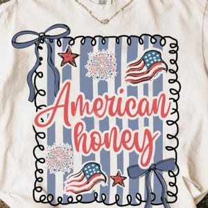 May include: White t-shirt featuring a patriotic design. The design includes the text "American honey" in red script, along with illustrations of the American flag, stars, fireworks, and blue bows. The design is framed with a black border.