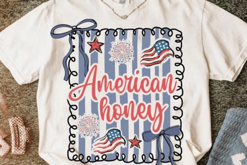 May include: White t-shirt featuring a patriotic design. The design includes the text "American honey" in red script, along with illustrations of the American flag, stars, fireworks, and blue bows. The design is framed with a black border.