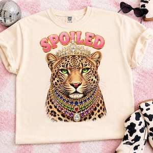 Op de afbeelding: Een crèmekleurig t-shirt met een afbeelding van een luipaard die een met juwelen bezette kroon en meerlaagse edelsteenkettingen draagt. Het woord SPOILED is in het roze boven de luipaard gedrukt. Het shirt heeft een ronde hals en korte mouwen.