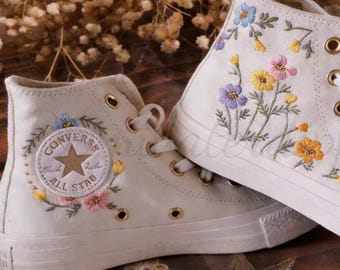 Könnte beinhalten: Cremefarbene High-Top-Sneaker mit gestickten Blumenmustern in Blau, Gelb und Rosa. Die Sneaker haben einen klassischen Converse-Logo-Patch und goldene Ösen. Die Schnürsenkel sind weiß und die Sohlen sind cremefarben.