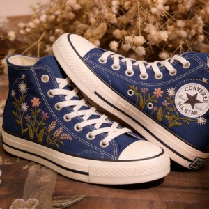 Könnte beinhalten: Ein Paar marineblaue High-Top-Sneaker mit weißen Schnürsenkeln und Sohlen. Die Sneaker haben gestickte Blumendesigns in Rosa, Gelb und Grün. Das Converse All Star-Logo ist auf der Seite sichtbar.
