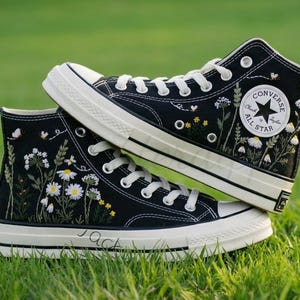 May include: Black high-top trainers with white laces and soles. The trainers are embroidered with white daisies, yellow flowers, and small butterflies. The Converse logo is visible on the side. The word "Jack" is written on the side of the sole.