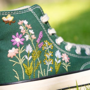 Könnte beinhalten: Ein hochgeschnittener Canvas-Sneaker in Dunkelgrün, verziert mit gestickten Wildblumen in Rosa, Gelb und Lila. Der Schuh hat weiße Schnürsenkel und eine weiße Gummisohle. Ein kleiner gestickter Schmetterling ist ebenfalls zu sehen.