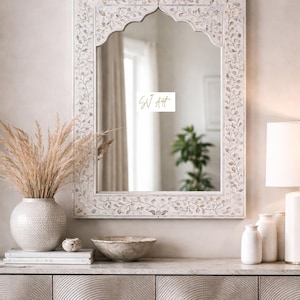 Puede incluir: Espejo blanco y ornamentado con un diseño floral alrededor del marco. El espejo es rectangular con una parte superior arqueada. Está sobre un mueble gris con un jarrón de flores secas, un cuenco y otros objetos decorativos.