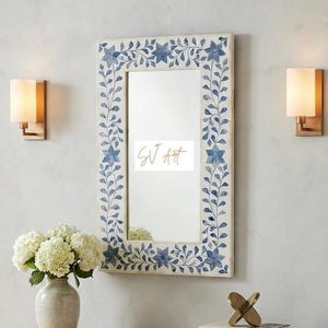 May include: A rectangular mirror with a white frame decorated with blue floral and leaf patterns. The mirror is above a light-coloured wooden console table. Two sconces flank the mirror, and a vase of white flowers sits on the table.